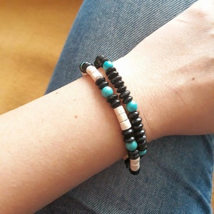 Puede incluir: Dos pulseras de cuentas apiladas en una muñeca. Una pulsera presenta cuentas negras y blancas, mientras que la otra tiene cuentas negras y turquesas. Las pulseras se usan sobre un fondo de jeans azules.