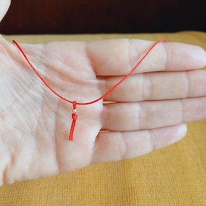 Peut inclure: Un collier de cordon rouge avec une petite perle dorée et un pompon. Le collier est sur une main.