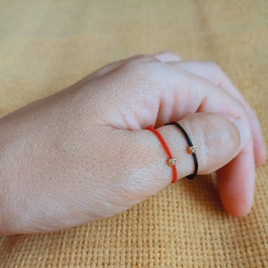 May include: Two red and black string bracelets with gold beads on a person's thumb.