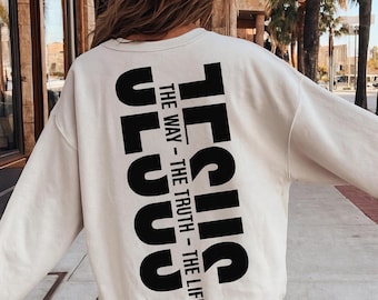 May include: A cream-coloured sweatshirt featuring the word "JESUS" printed vertically in large, bold, black letters on the back. Below the word are the phrases "THE WAY - THE TRUTH - THE LIFE" in a smaller, black font. The sweatshirt has long sleeves.