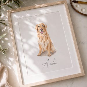 May include: A framed watercolor painting of a golden retriever dog. The dog is depicted sitting with its tongue out. The dog's name, "Archie", is written in cursive below the dog. The frame is light-colored wood.