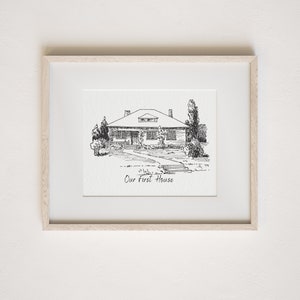 Op de afbeelding: Een zwart-witte lijntekening van een huis met een voorporch en een pad dat naar de voordeur leidt. De tekening is ingelijst in een licht houten lijst en de tekst "Our First House" staat onder de tekening.
