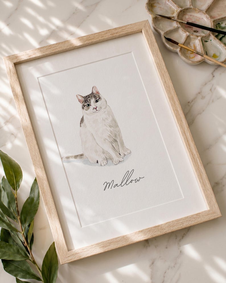 May include: A framed watercolor illustration of a white cat with grey tabby markings on its head and tail. The cat is sitting in a centered position. Below the cat, the name Mallow is written in a black cursive script on the white paper.