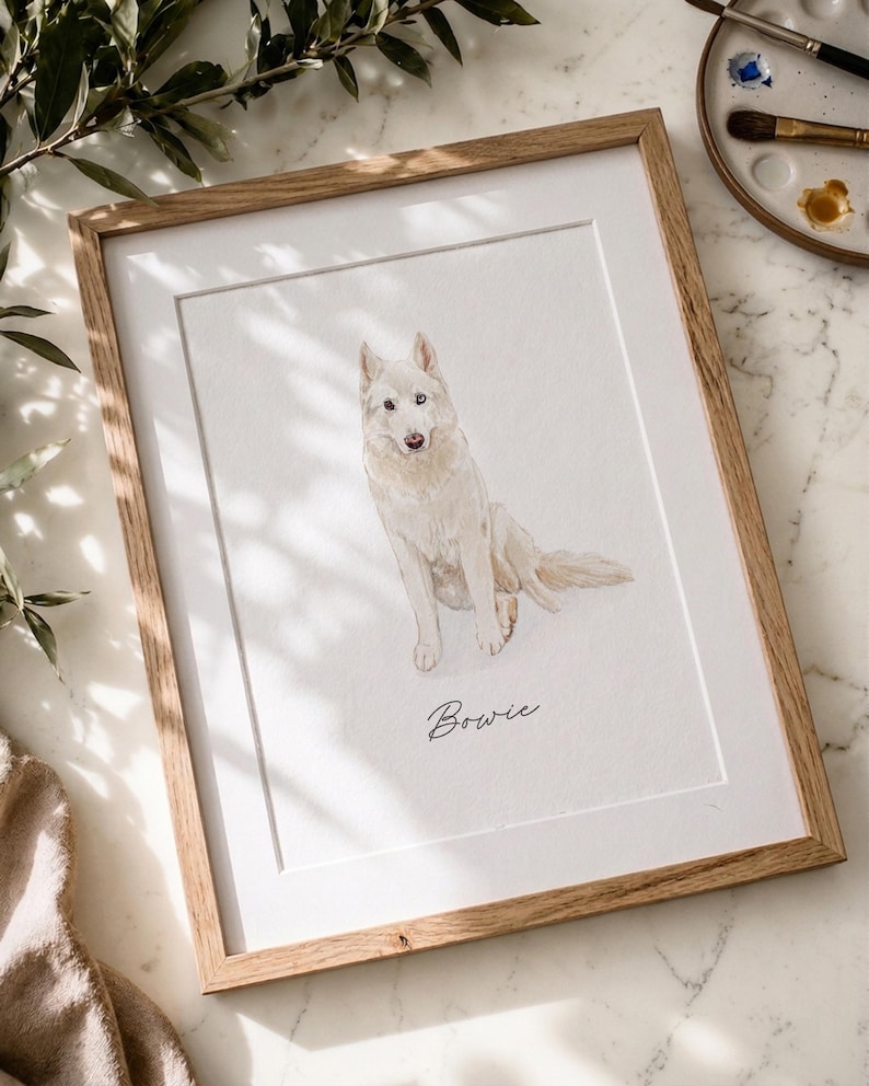 May include: A framed watercolor portrait of a white dog sitting down, featuring the name Bowie written in black cursive script below the illustration. The artwork is encased in a light-toned wooden frame with a white mat border.