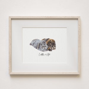 May include: A watercolour painting of two rabbits, one grey and one brown, cuddling together. The painting is framed in a light wood frame and has the text "Cuddles & Kitten" written below the rabbits.