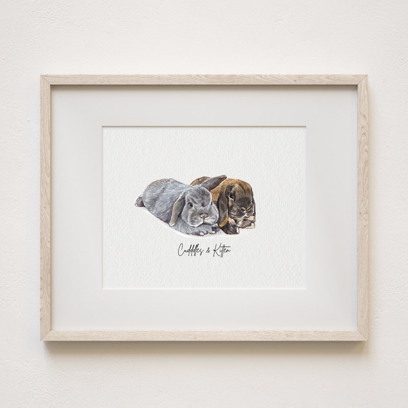 May include: A watercolour painting of two rabbits, one grey and one brown, cuddling together. The painting is framed in a light wood frame and has the text "Cuddles & Kitten" written below the rabbits.