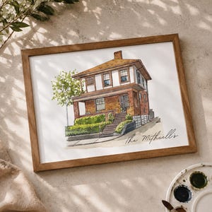 May include: Watercolor painting of a two-story brick house with a brown roof, framed in a wooden frame. The house has white trim and a small tree to the left. The name "The Mitchell's" is written in script.