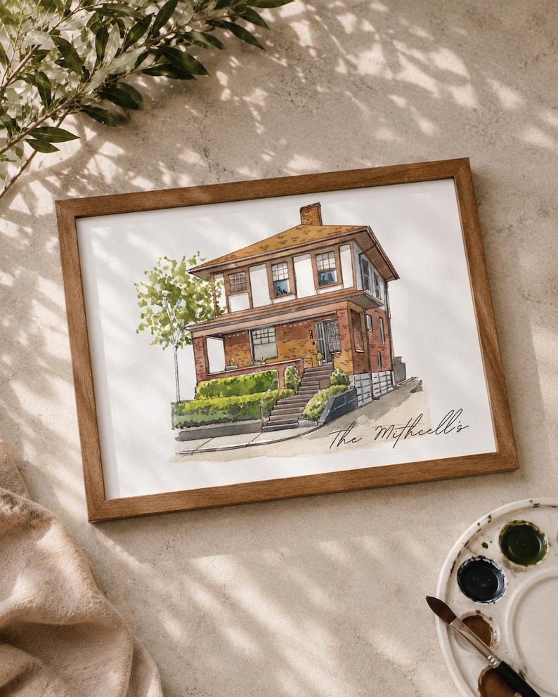 May include: Watercolor painting of a two-story brick house with a brown roof, framed in a wooden frame. The house has white trim and a small tree to the left. The name "The Mitchell's" is written in script.