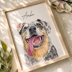 Pode incluir: Retrato em aquarela de um cão chamado Archie, emoldurado em madeira clara. O cão tem uma pelagem preta, cinza e castanha, com uma língua rosa. A obra de arte está em um fundo branco.