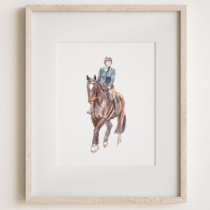 May include: A framed watercolor illustration of a brown horse with a white blaze on its face, ridden by a person wearing a dark blue equestrian jacket, tan breeches, and a black helmet. The artwork is encased in a light-toned wooden frame.