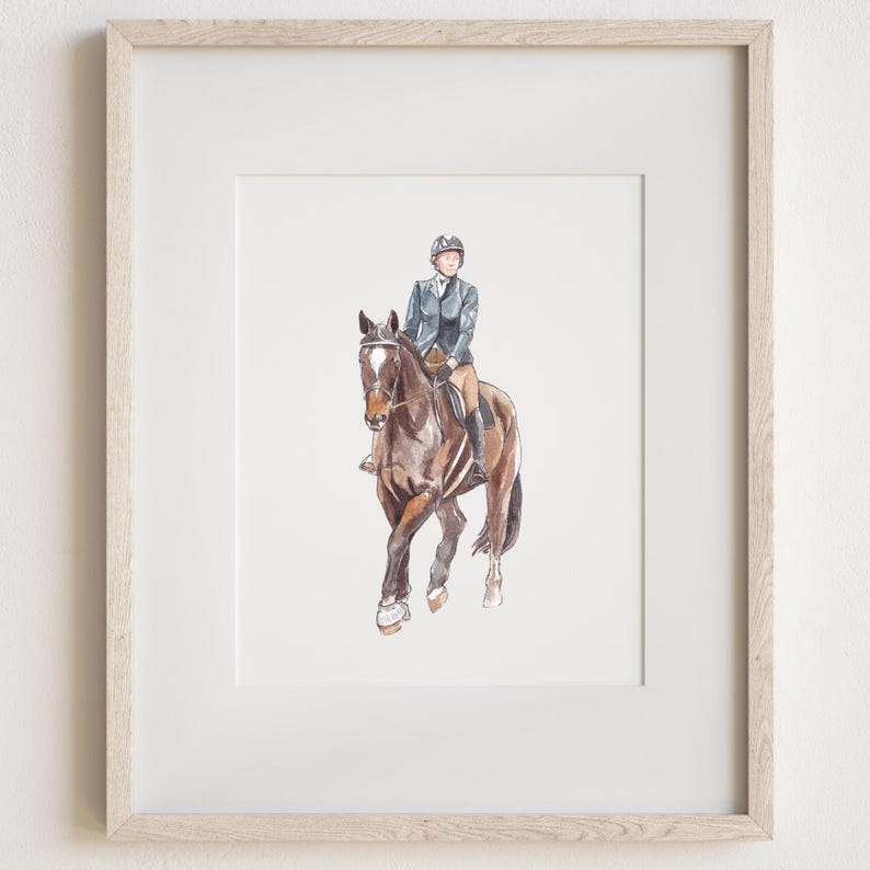 May include: A framed watercolor illustration of a brown horse with a white blaze on its face, ridden by a person wearing a dark blue equestrian jacket, tan breeches, and a black helmet. The artwork is encased in a light-toned wooden frame.