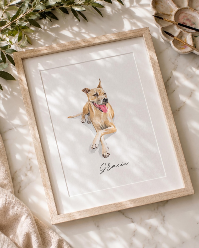 May include: A framed watercolor portrait of a tan dog with its tongue out, lying down with crossed front paws. The name Gracie is written in black cursive script below the dog. The artwork is enclosed in a light-colored wooden frame.