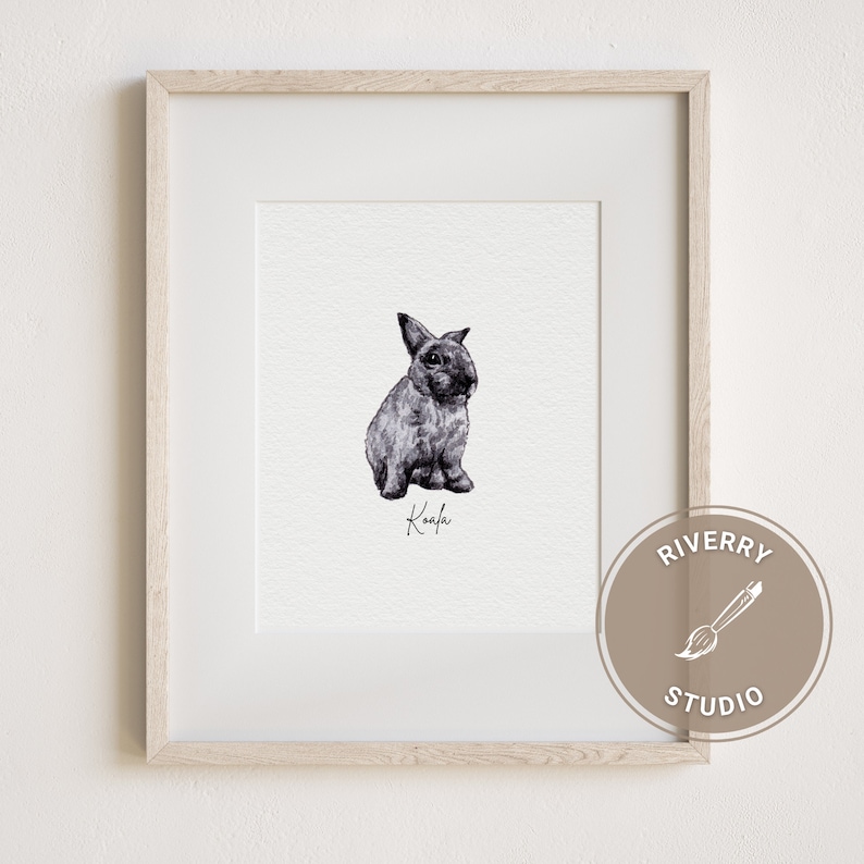 May include: A black and white watercolour illustration of a rabbit sitting on a white background. The rabbit is named "Koala" in cursive handwriting. The image is framed in a light wood frame.