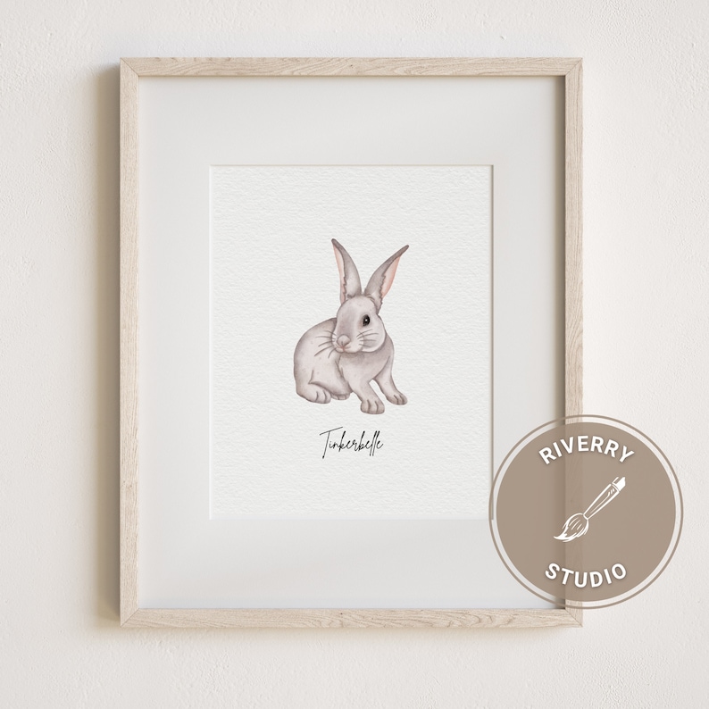 May include: A watercolour illustration of a grey rabbit with pink ears and nose. The rabbit is sitting on a white background with the name "Tinkerbelle" written below it. The image is framed in a light wood frame with a brown circle in the bottom right corner that says "Riverry Studio" with a paintbrush icon.