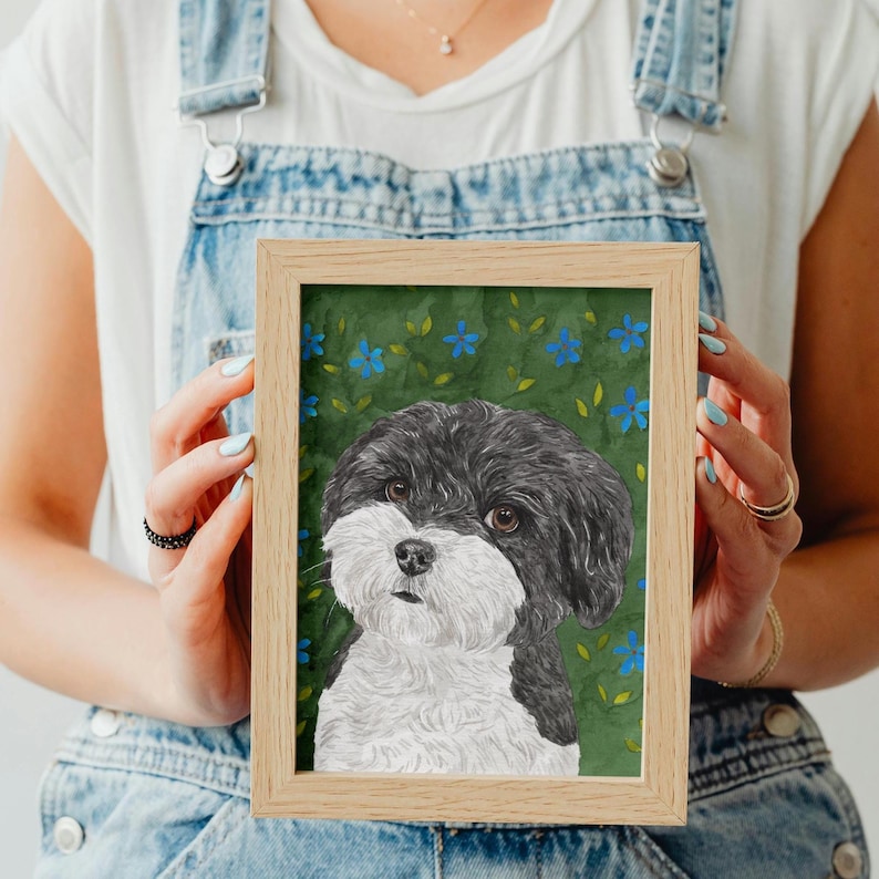 以下が含まれることがあります： ふわふわした毛並みの白黒の犬の肖像画が、小さな青い花と緑の葉が描かれたダークグリーンの背景に飾られています。このアートワークは、シンプルで清潔感のある縁取りの明るい木製フレームに収められています。