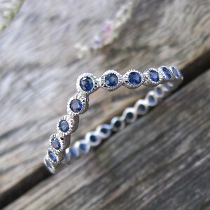 May include: A silver ring with a delicate design, featuring a series of small, round, blue gemstones set in a scalloped bezel. The ring is set against a rustic wooden surface, with a few sprigs of dried flowers in the background.