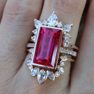 Pode incluir: Um anel de ouro com uma grande gema retangular vermelha cercada por gemas menores e transparentes. O anel tem um design floral delicado.