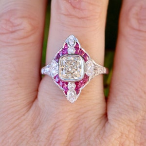 以下が含まれることがあります： 正方形のダイヤモンドが、小さなダイヤモンドと赤いルビーで囲まれた銀の指輪