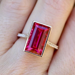 Könnte beinhalten: Ein rechteckiger Ring mit einem rubinroten Edelstein und einer goldfarbenen Lünettenfassung. Das Ringband ist mit kleinen, funkelnden Steinen verziert. Der Ring wird auf einer Hand mit verschwommenem grünem Hintergrund gezeigt.