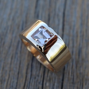 May include: A gold ring with a square-shaped clear gemstone set in a raised bezel. The ring has a wide band with a smooth, rounded surface.