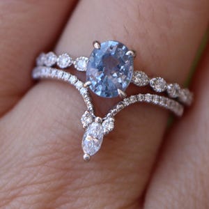 May include: A close-up of a two-piece engagement ring set. The primary ring features a light blue oval-cut gemstone. The band is adorned with small, clear gemstones. The second ring has a curved design with small, clear gemstones.