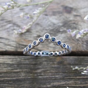 May include: A silver ring with a curved design, set with small, round blue gemstones. The ring is on a wooden surface, with sprigs of purple flowers in the background. The ring's design is a series of bezel-set stones.