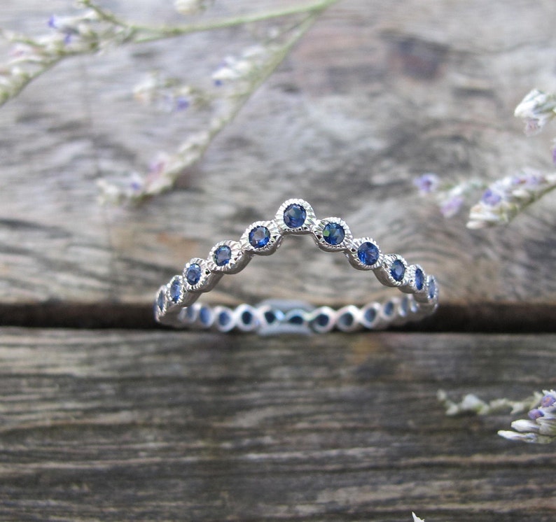 May include: A silver ring with a curved design, set with small, round blue gemstones. The ring is on a wooden surface, with sprigs of purple flowers in the background. The ring's design is a series of bezel-set stones.