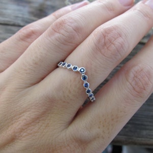 May include: A silver ring with a curved design, set with small, round, blue gemstones. The ring is shown on a person's finger, with the gemstones arranged in a delicate pattern. The ring's design is elegant and suitable for everyday wear.