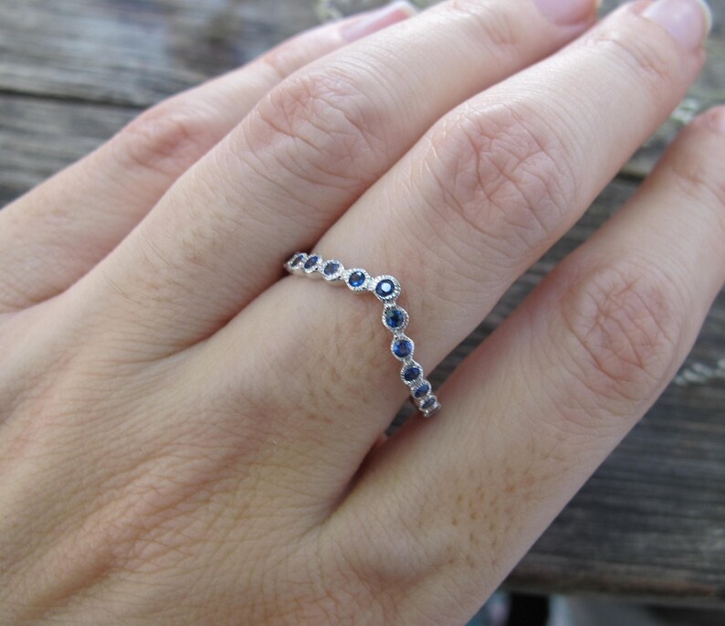 May include: A silver ring with a curved design, set with small, round, blue gemstones. The ring is shown on a person's finger, with the gemstones arranged in a delicate pattern. The ring's design is elegant and suitable for everyday wear.
