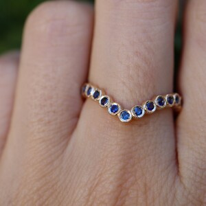 May include: A gold-colored ring with a V-shaped design. The ring is adorned with a series of small, round, blue gemstones. The gemstones are set closely together, creating a continuous line of color. The ring is a piece of jewelry.