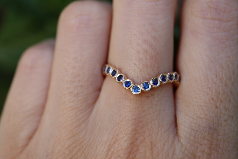 May include: A gold-colored ring with a V-shaped design. The ring is adorned with a series of small, round, blue gemstones. The gemstones are set closely together, creating a continuous line of color. The ring is a piece of jewelry.