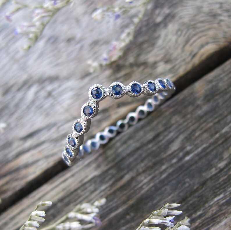 May include: A silver ring with a delicate design, featuring a series of small, round, blue gemstones set in a scalloped bezel. The ring is set against a rustic wooden surface, with a few sprigs of dried flowers in the background.