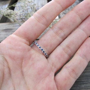May include: A silver ring with a continuous line of small, round, dark blue gemstones. The ring is set against a blurred background of a hand and wooden surface, with hints of dried flowers.