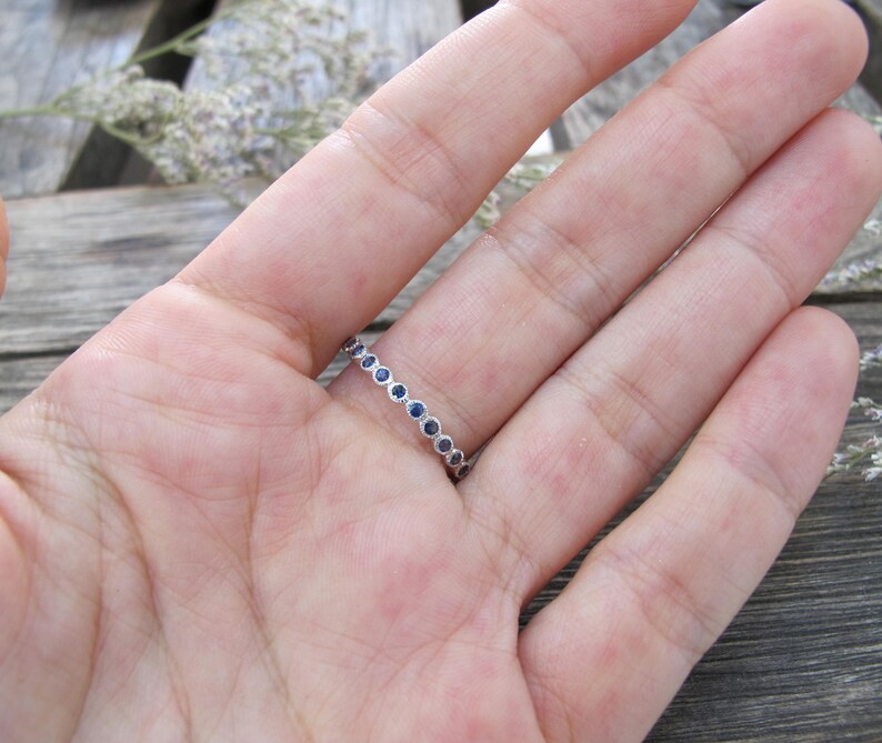 May include: A silver ring with a continuous line of small, round, dark blue gemstones. The ring is set against a blurred background of a hand and wooden surface, with hints of dried flowers.