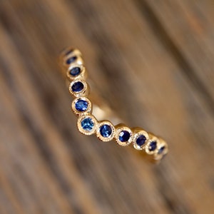 May include: A gold-colored ring with a V-shaped design. The ring is adorned with a series of small, round, blue gemstones. The gemstones are set closely together, creating a continuous line of color. The ring is set against a blurred wooden background.