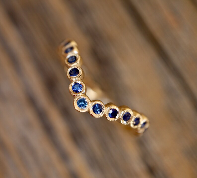 May include: A gold-colored ring with a V-shaped design. The ring is adorned with a series of small, round, blue gemstones. The gemstones are set closely together, creating a continuous line of color. The ring is set against a blurred wooden background.