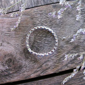 May include: A silver ring with a continuous band of small, round, blue gemstones. The ring is set against a weathered wooden surface, with sprigs of dried purple flowers in the background. The ring's design is simple and elegant.