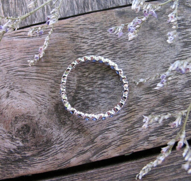 May include: A silver ring with a continuous band of small, round, blue gemstones. The ring is set against a weathered wooden surface, with sprigs of dried purple flowers in the background. The ring's design is simple and elegant.