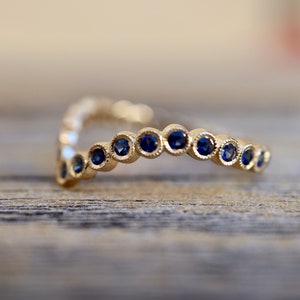 May include: A gold-colored ring with a V-shape design. The ring is adorned with a series of small, round, dark blue gemstones. The gemstones are evenly spaced along the ring's surface, creating a continuous pattern. The ring is set against a blurred wooden background.
