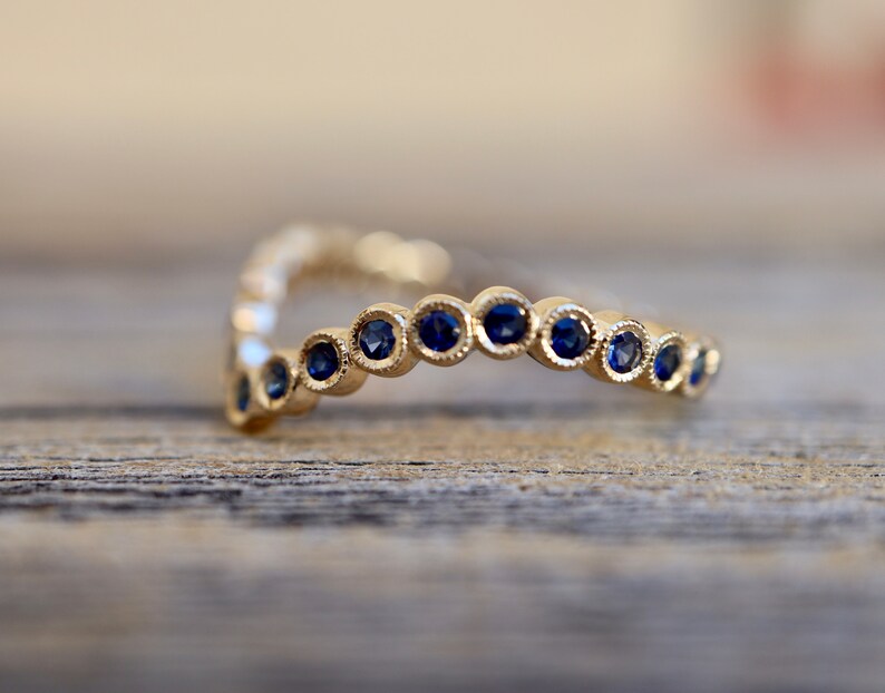 May include: A gold-colored ring with a V-shape design. The ring is adorned with a series of small, round, dark blue gemstones. The gemstones are evenly spaced along the ring's surface, creating a continuous pattern. The ring is set against a blurred wooden background.