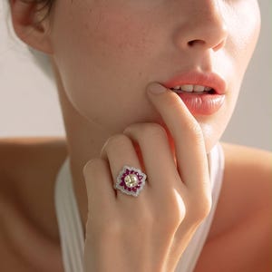Puede incluir: Un anillo de plata con una piedra preciosa central amarilla de corte cojín, rodeada por un halo de pequeñas piedras rojas vibrantes. El engaste está acentuado con diminutas piedras transparentes en pavé dispuestas en un patrón geométrico floral festoneado.