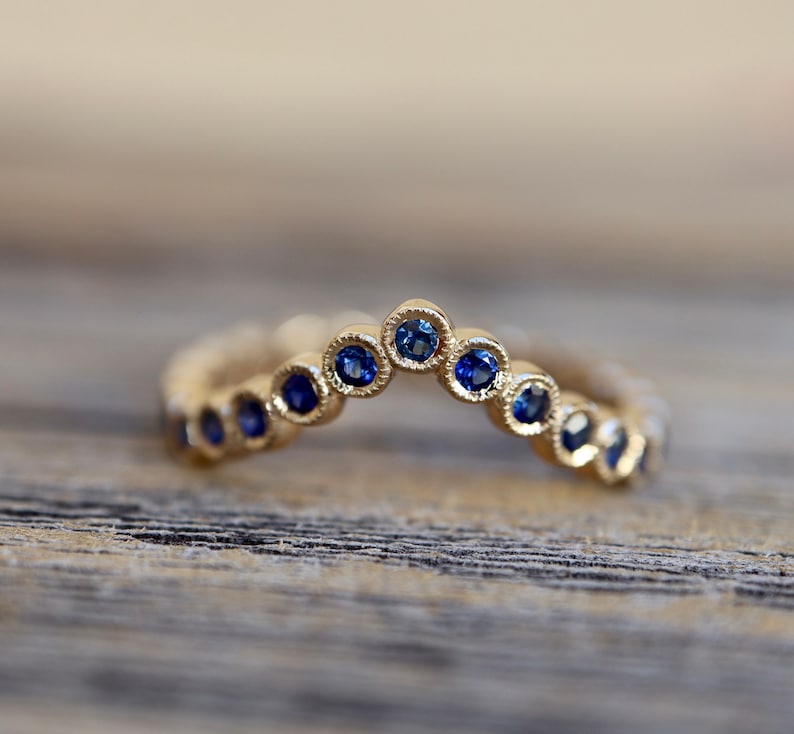 May include: A gold-colored ring with a curved design, featuring a series of small, round, blue gemstones. The ring is set against a blurred wooden background, highlighting the jewelry's intricate details and craftsmanship.