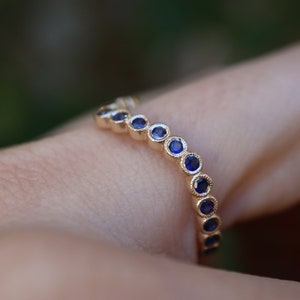 May include: A gold ring with a series of small, round, deep blue gemstones set in a row. The ring is on a finger, with a blurred green background.