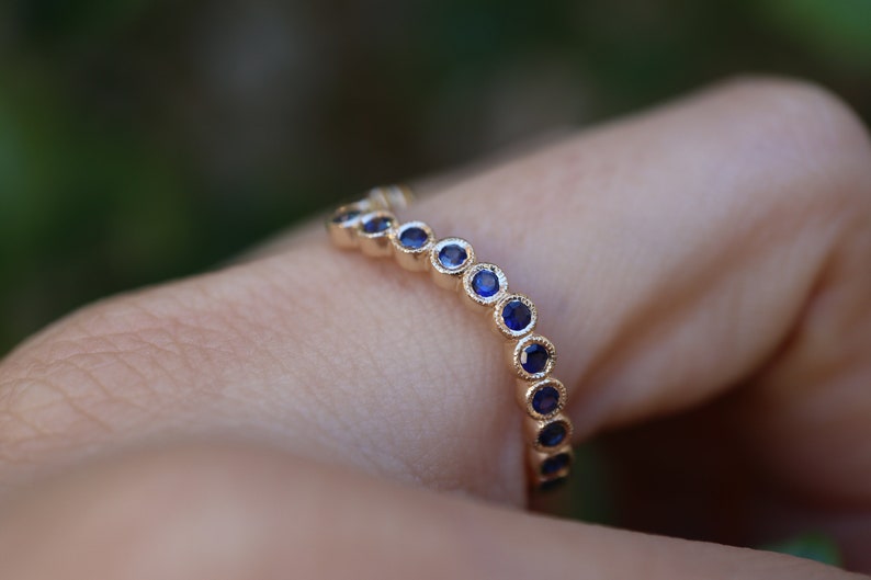 May include: A gold ring with a series of small, round, deep blue gemstones set in a row. The ring is on a finger, with a blurred green background.