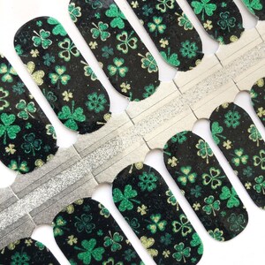 May include: A sheet of nail polish wraps with a black background and a green shamrock pattern. The shamrocks are outlined in gold and have a glitter finish.