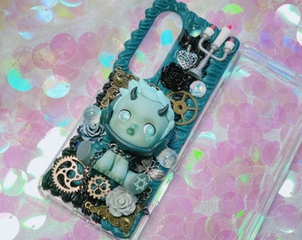 Può includere: Una custodia per telefono blu-verde con un personaggio scolpito in 3D blu e bianco con corna, una catena e vari abbellimenti, tra cui ingranaggi, fiori e un candelabro.
