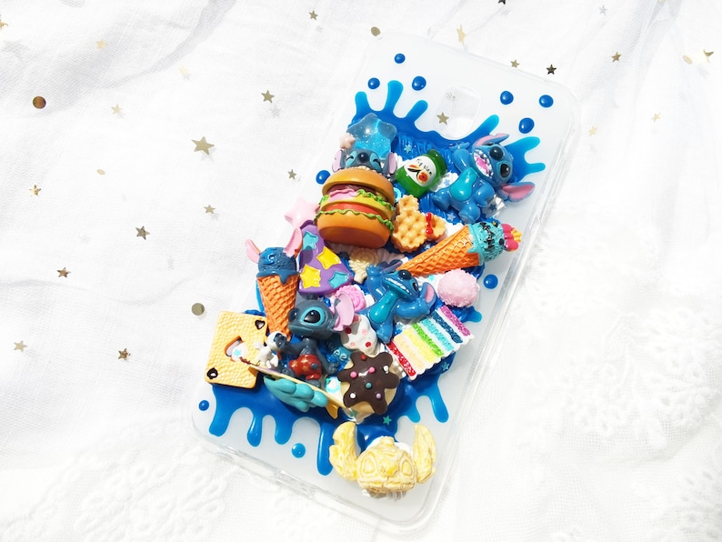 Op de afbeelding: Een transparante telefoonhoes met een blauw en wit splatterontwerp. De hoes is versierd met verschillende 3D-charms, waaronder een blauw cartoonfiguur, een hamburger, ijshoorntjes en andere etenswaren.