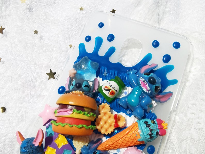 Op de afbeelding: Een transparante telefoonhoes met een blauw en wit ontwerp met een cartoonfiguur, Stitch, uit de Disney-film Lilo & Stitch. De hoes bevat ook een hamburger, een ijsje en een wafel.
