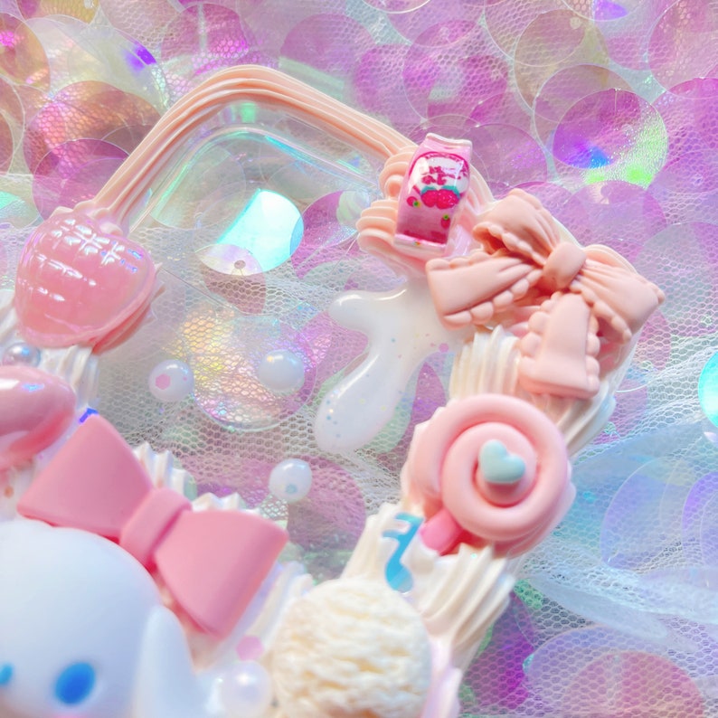 May include: A pink and white phone case with a variety of cute decorations, including a bow, a lollipop, and a heart. The case is decorated with a white and pink swirl design.