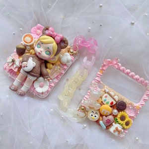 May include: Two decorative phone cases with a whimsical design. One features a doll figure in a brown sweater, the other has a handle and is decorated with sweets and a sunflower. Both cases are pink and white with pearl accents.